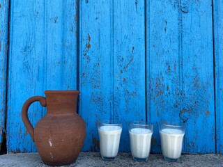 Brown old clay jug and transparent glasses with milk stand on gray concrete on a background of blue...