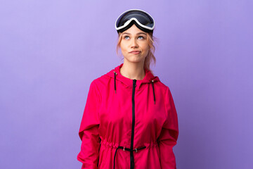 Skier teenager girl with snowboarding glasses over isolated purple background and looking up