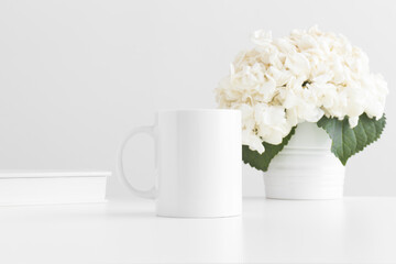 White mug mockup with a book and a hortensia in a pot on a white table.