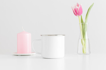 Enamel mug mockup with pink tulips in a vase and candle on a white table.