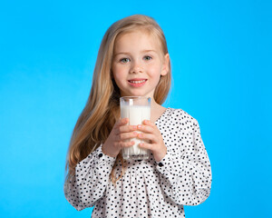 Beautiful European blonde girl with a glass of milk in her hands. Baby on a blue background for advertising milk.