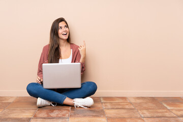 Teenager student girl sitting on the floor with a laptop pointing up and surprised