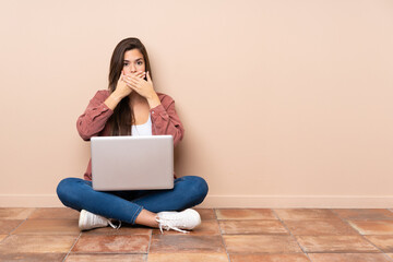 Teenager student girl sitting on the floor with a laptop covering mouth with hands