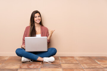 Teenager student girl sitting on the floor with a laptop pointing to the side to present a product
