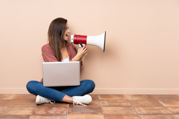 Teenager student girl sitting on the floor with a laptop shouting through a megaphone
