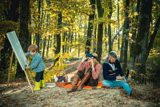 Family vacation at autumnal golden forest. Different types of activities: father do his business using laptop, mother has fun with augmented reality glasses and their little son paint.