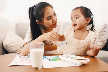 Young Asian mother playing with her cute daughter with glass of milk and watercolor painting work on the table at home. Family lifestyle mother and child activity concept