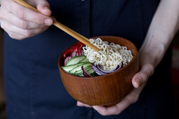 The female hand holds chopsticks with noodles, eats noodles and vegetables from a wooden bowl. Thai and asian sharp food, beautiful serve. Light photo, minimalism.
