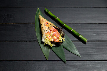 Japanese Sushi Temaki Hand Roll with salmon, rice, Tobiko roe eggs, avocado and salad mix in Mamenori seaweed. Close up of Pan Asian traditional dish on bamboo leaves near bamboo stick on wooden board