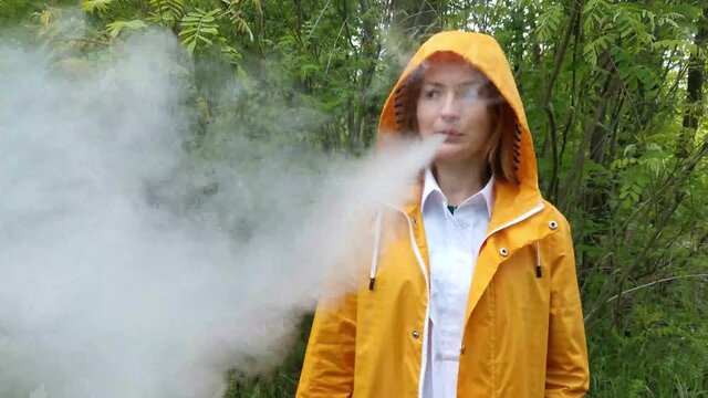 Young girl in a jacket with a hood smoking with steam en a background of green foliage