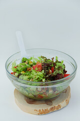 fresh vegetable salad tomatoes, cucumbers, lettuce leaves, pine nuts, walnuts in a glass plate with a white spoon on a wooden stand on a white background