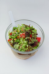 fresh vegetable salad tomatoes, cucumbers, lettuce leaves, pine nuts, walnuts in a glass plate with a white spoon on a wooden stand on a white background