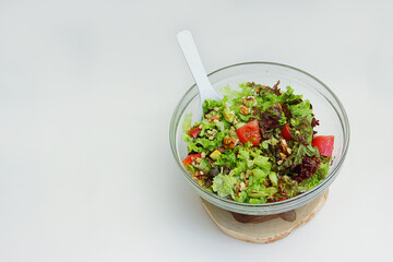 fresh vegetable salad tomatoes, cucumbers, lettuce leaves, pine nuts, walnuts in a glass plate with a white spoon on a wooden stand on a white background and there is a place for the inscription