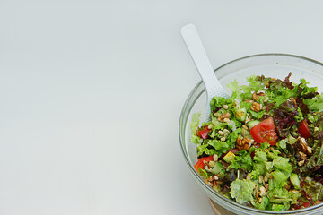 fresh vegetable salad tomatoes, cucumbers, lettuce leaves, pine nuts, walnuts in a glass plate with a white spoon on a wooden stand on a white background
