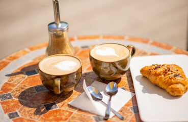 Two ceramic cappuccino cups and a croissant on a mosaic cafe table, on a Sunny summer day. Concept for meetings with friends, summer walks. Close-up, selective focus, horizontal position