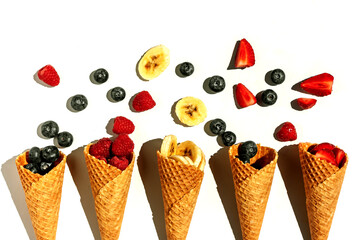 Various summer fruits in ice cream cones on white background. Strawberry, blueberry, raspberry, banana slices in waffle cups. Summer sweet menu concept flat lay.