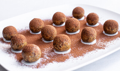 Useful, delicious energy balls lie on a white dish, sprinkled with cocoa. A nutritious home-made product made from oat flakes, nuts and seeds. Close-up, selective focus.