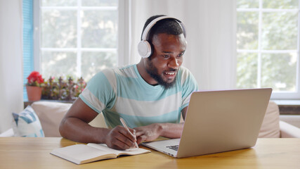 Portrait of young cheerful afro-american guy in headphones watching online courses on laptop and writing down notes. Remote education and e-learning concept