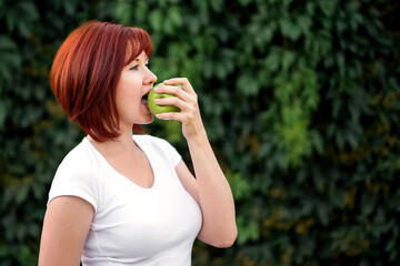 Portrait of middle-aged woman eating big green apple outdoors. Blurred green leaves in background....