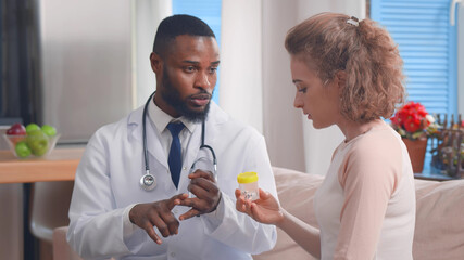 Young african male doctor with bottle of pills in hands treating patient at home