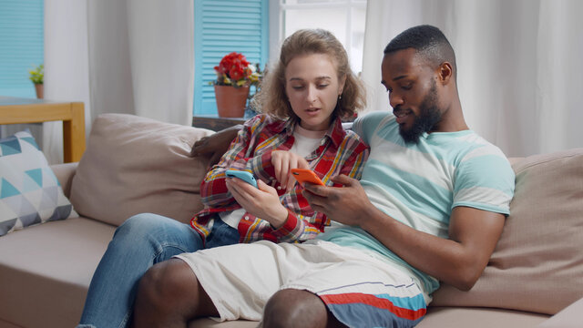 Cheerful diverse man an woman in love using phones sitting on sofa