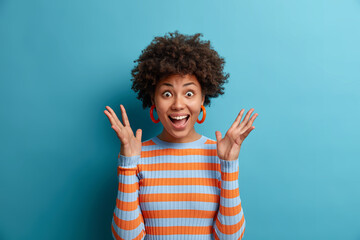 Portrait of delighted cheerful ethnic woman reacts on awesome gift, raises palms and stares with eyes full of happiness, cannot believe in her great success, dressed in casual wear, poses indoor