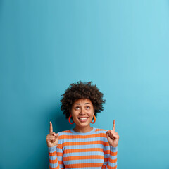 Cheerful African American woman looks up and points upwards with broad smile, recommends awesome product, suggests click banner, dressed casually, isolated on blue background shows advertisement promo - obrazy, fototapety, plakaty