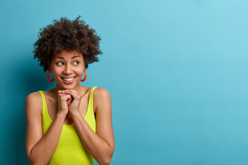 Horizontal studio shot of cheerful African American woman keeps hands under chin and looks...