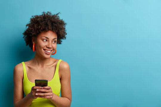 Horizontal shot of pretty smiling woman types on smart phone, waits for call, looks away with glad expression, texting in social media, uses mobile network service, isolated on blue background