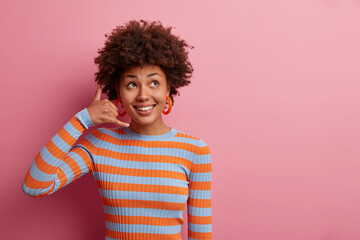 Give me call sometimes. Cheerful smiling ethnic woman shapes hand like phone, looks gladfully...
