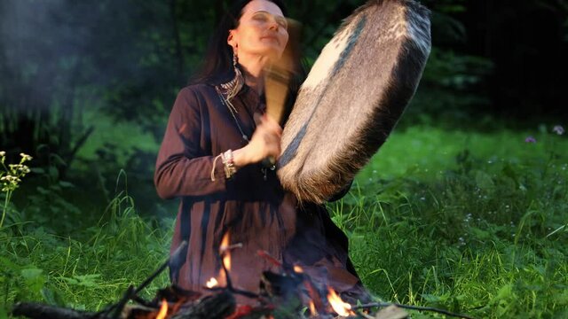 Female shaman performs a shamanic rite with a tambourine while in a trance by the fire in the forest
