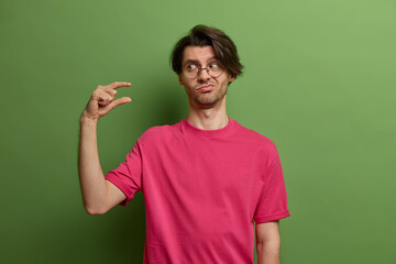 Displeased man asks some more time, shows small size or little bit gesture, needs minimum amount, measures not big scale, dressed casually, isolated on green background. Body language concept