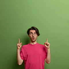 Alarmed shocked man points index fingers up and shows something disturbing, tells surprising news demonstrates awesome discount or unexpected sale offer poses against green background free space above