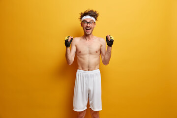 Funny positive skinny man raises hands with clenched fists, feels very happy and motivated, wears...