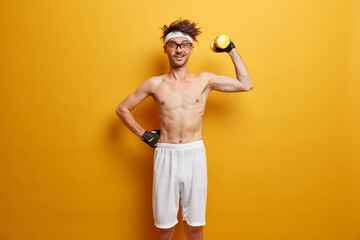 Indoor shot of positive man holds heavy sport equipment, enjoys workout in own gym, has skinny body, wants to be healthy and fit, wears transparent glasses, sport gloves, poses over yellow wall