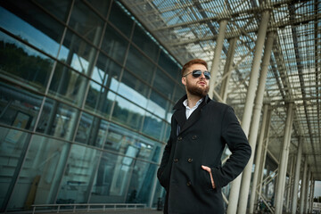 Success. Stylish young bearded man in coat, sunglasses businessman standing near the business center, looking far. With hands pockets, pensive and concentrated. Confident look of