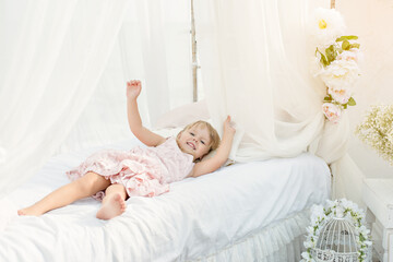 Obraz na płótnie Canvas Beautiful little blonde girl with blue eyes in a pink tender dress barefoot, lies on white sheets with pillows of a bed with chiffon canopies on a background of blue sea. Happy childhood and vacation