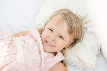 Fototapeta na wymiar Beautiful little blonde girl with blue eyes. A close up portrait, the baby lies on white sheets with pillows of a bed with chiffon canopies against the blue sea. Happy childhood and summer vacation