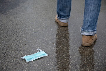 Medical face mask discarded on the wet asphalt, man's legs walk  away, reckless behavior against the risk of coronavirus infection after the lockdown, copy space