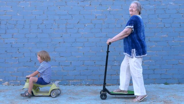 Funny blond boy in plaid shirt riding bicycle with his beloved grandmother. child teaches an elderly granny to ride. pensioner is smartly dressed and rolls on a large stretched scooter