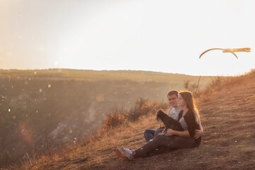 Naklejka na ściany i meble Happy couple in love are resting on the mountainside with a black Schnauzer, sitting on the ground, hugging a dog, watching bright paragliders fly in the blue sky, admire the sunset, travel with a dog