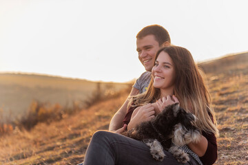 Happy couple in love are resting on the mountainside with a black Schnauzer, sitting on the ground, hugging a dog, watching bright paragliders fly in the blue sky, admire the sunset, travel with a dog