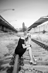 Fototapeta na wymiar A sad and lonely girl with long hair blonde in a black short dress with long legs sits on the rails of railway tracks. The pain of loss, loss, broken heart, disappointment after parting with loved one