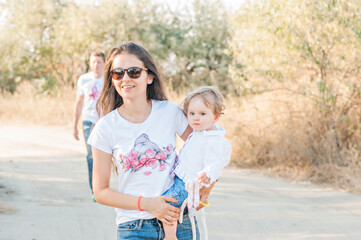 Fototapeta na wymiar Modern stylish mother in jeans and a white T-shirt, holds a little daughter in her arms, walk in the park in the fall. Young brunette woman in black glasses smiling, happy motherhood walking in nature