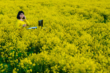 Young woman freelancer in a field among yellow flowers at a table with and a computer.