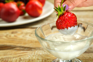 A female hand holds a strawberry in cream with a cup of whipped cream.
