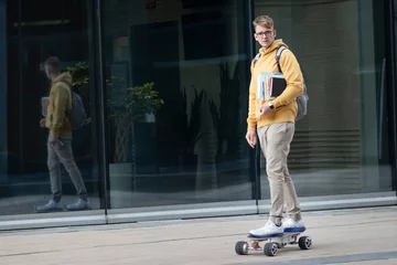 Poster Handsome guy, young man, hipster, student or pupil in glasses on his face riding on electric urban modern skateboard with backpack, books and textbooks. Eco transport, technology concept. © Евгений Шемякин