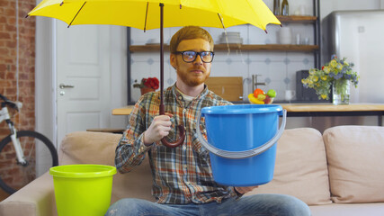 Man with bucket under umbrella suffering from leaking at home. Waiting for repairman © TommyStockProject