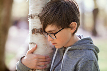 A sad boy in a park is holding a birch tree