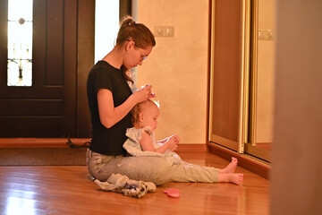 Mom makes a hairstyle to her little daughter in the hallway, the sun breaks through the front door, creating backlight. Warm toned